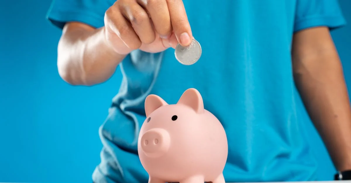 Gros plan sur la main d'un adulte déposant une pièce dans une tirelire, symbolisant l'épargne et l'investissement.