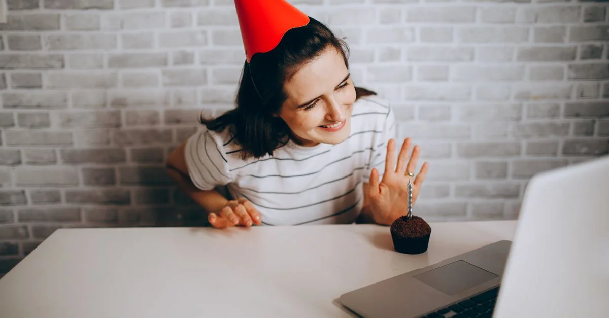 Femme avec un chapeau de fête saluant un ordinateur portable et un cupcake, célébrant un anniversaire virtuel.