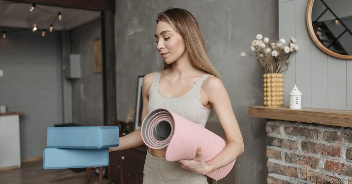 Femme en tenue de sport tenant un tapis de yoga et des blocs dans un cadre intérieur confortable.