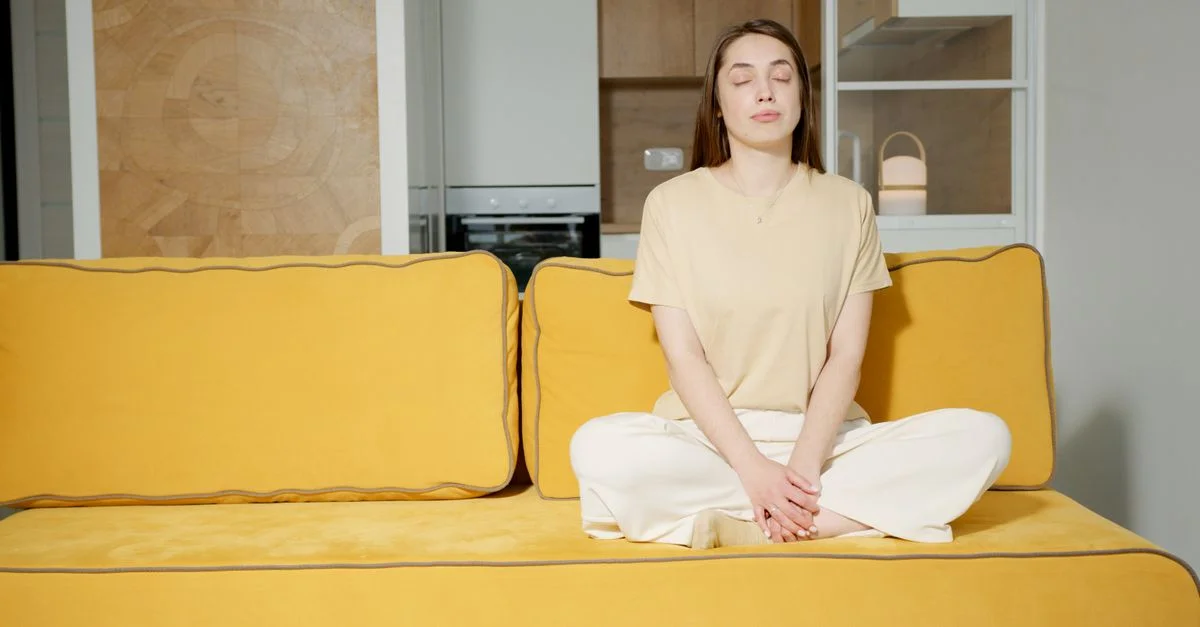 Femme détendue méditant les yeux fermés sur un canapé jaune moderne dans un cadre domestique minimaliste.