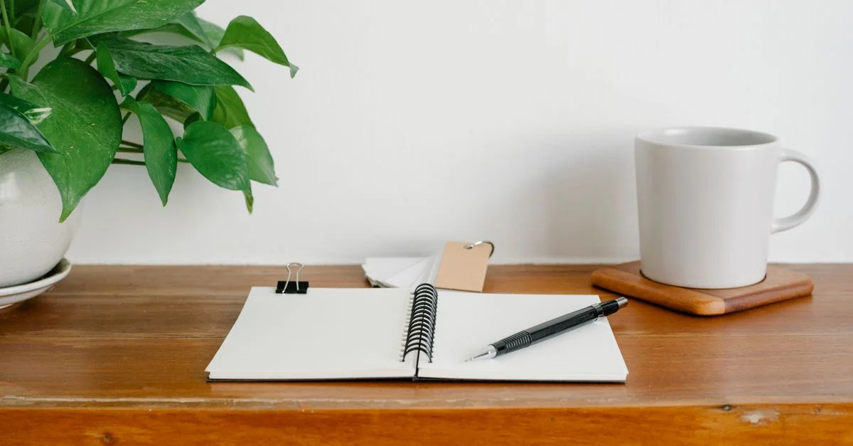 Un espace de travail propre et organisé avec un bloc-notes, un stylo, une tasse et une plante en pot sur un bureau en bois.